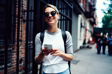 Fototapeta premium Happy hipster girl sending message via smartphone enjoying leisure strolling ,smiling female tourist in sunglasses satisfied with internet connection in roaming chatting online while sightseeing.