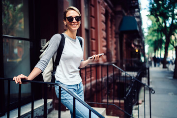 Half length portrait of cheerful young woman in sunglasses standing on stairs holding smartphone,positive hipster girl in casual wear looking at camera sending message via mobile phone on street