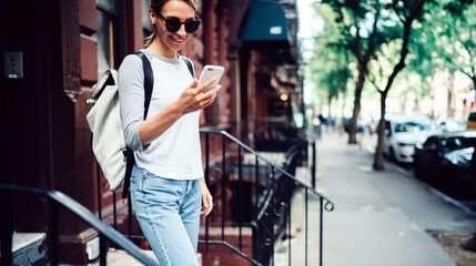Cheerful hipster girl getting message on mobile phone standing on stairs on city street,positive casually dressed young woman blogging in networks using smartphone spending free time outdoors