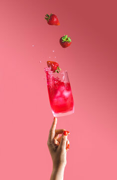 Woman hand support fly glass of strawberry drink with splash, juice strawberries falling in glass. Summer art food concept on pink background
