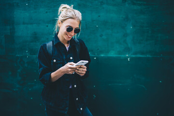 Smiling good looking young woman typing message on smartphone standing near promotional background,...