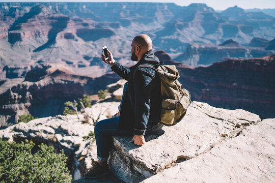 Male traveler with backpack using smartphone camera for taking picture of beautiful landscape of Grand Canyon National Park, hipster guy wanderlust checking good mobile connection sitting on hill peak