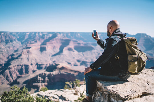 Rear view of male traveler with rucksack shooting video on mobile phone for share in social media while having exploring wanderlust tour, millennial hipster guy posing for selfie on cellular camera - Powered by Adobe