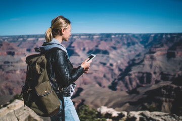 Positive hipster girl wanderlust with smartphone good connection for sending data during weekends in Grand canyon, smiling female hiker looking at breathtaking nature landscape holding cellphone .