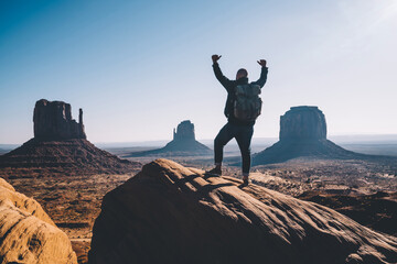 Happy male traveler feeling free standing on top of high rock ver Monument valley, back view of hipster guy excited with successful journey lifestyle raising hands up looking at sandstones in desert