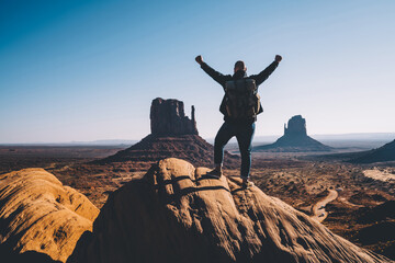 Silhouette back view of excited male traveler raising hands up standing on rock over desert with...