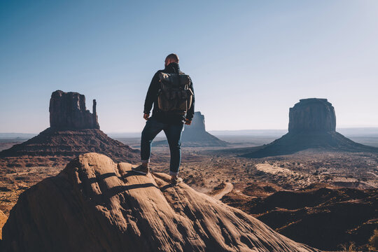 Back view of man with backpack standing on rock looking at high beautiful scenery view in dry desert lands of Monument Valley,male traveler wanderlust getting to destination exploring wild environment