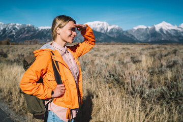 Young female explorer observing breathtaking nature view enjoying hiking tour with backpack, cheerful girl wanderlust looking at scenery of mountains landscape spending leisure on discovering.