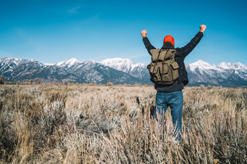 Back view of male traveler feeling freedom raising arms and celebrating victory, happy hipster guy with backpack excited from spring holidays in mountains during hiking tour exploring wild nature