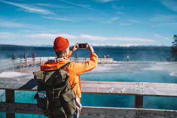 Back view of female traveler using smartphone camera for taking picture of hot springs, hipster girl with backpack making photo getting to touristic landmark in Yellowstone during american trip