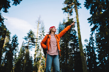 Young female traveler in colourful jacket pointing on high tree in forest hiking trails in wild...