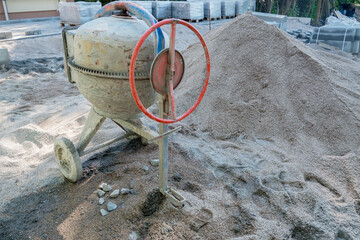 A small metal concrete mixer on wheels with a rounded body, electric drive and a red wheel of tipping the finished cement mortar on a jagged rim near a pile of gray sand on a construction site.
