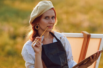  a girl artist with long red hair draws on an easel with a brush against the background of the sunset