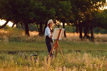  a girl artist with long red hair draws on an easel with a brush against the background of the sunset