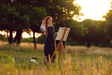  a girl artist with long red hair draws on an easel with a brush against the background of the sunset