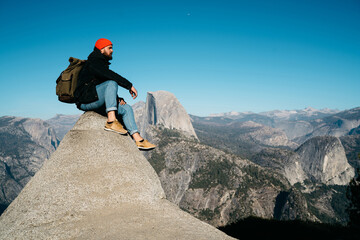 Young male traveler sitting on high rocky cliff over abys looking at breathtaking scenery of nature spending weekends actively,hipster guy enjoying wanderlust adventure resting on mountain top.
