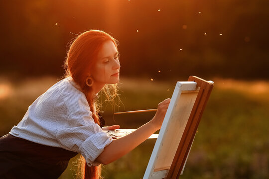  a girl artist with long red hair draws on an easel with a brush against the background of the sunset