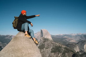 Foto op Aluminium Male traveler with touristic backpack sitting on high rock cliff pointing on landscape of Yosemite national park,hipster guy enjoying wanderlust trip to mountain observing wild land for hiking. © BullRun