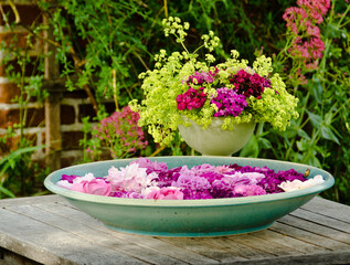 A large bvowl of crockery filled with water and many swimming leafs of pink flowers on a wooden table in front of a beautiful flower bouquet in a garden