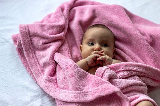 Photo of pretty chubby baby girl portrait, in diaper pants,lying in bed wrapped in soft pink blanket.Looking up.Caucasian baby girl looking at camera.Portrait of lovely baby wrapped in pink blanket.