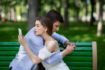 Gadget addicted couple using mobile phones while embracing on bench at park