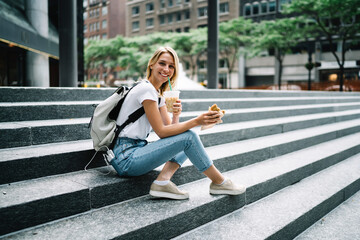 Portrait of cheerful female tourist sitting on stairs having meal tasting fast food and beverage,smiling hipster girl looking at camera having lunch break outdoors eating snack and drinking coffee