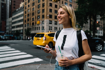 Cheerful young female tourist walking on city street passing cars and crosswalk enjoying big city,smiling hipster girl excited with fast taxi service calling cab via application on smartphone.