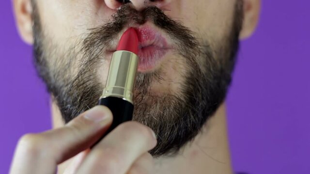 A young guy with a beard paints his lips with red lipstick. Close-up of a bearded man, he painted lips with bright lipstick. A bearded man puts red lipstick on his lips, smiles and smacks his lips.