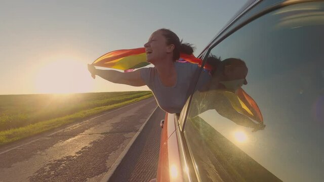 Happy woman with flag of LGBT people in car window traveling on road at sunset.