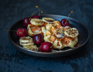 Homemade cottage cheese and banana waffles with mugs of bananas and cherry berries and grated chocolate on a black plate
