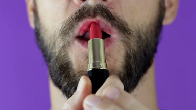 A young guy with a beard paints his lips with red lipstick. Close-up of a bearded man, he painted lips with bright lipstick. A bearded man puts red lipstick on his lips, smiles and smacks his lips.