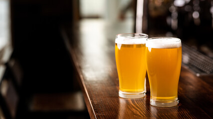 Hangouts with friends in pub and beer set. Two glasses of light beer with foam on wooden bar counter in daylight in interior of pub