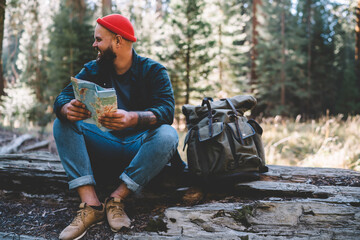 Happy male traveller smiling in nature environment while resting on wooden log with location paper map in hands, positive hipster guy with rucksack feeling good from spending weekend in wild forest