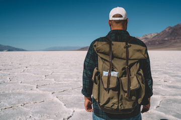 Back view of male traveler with rucksack having adventure in arid wildlands of Badwater during daytime, man walking on cracked surface of dry lake exploring landscape with mountains and low valley