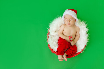 a sleeping child in a Santa costume lies with a candy in his hands on a green isolated background, waiting for the new year and Christmas, space for text