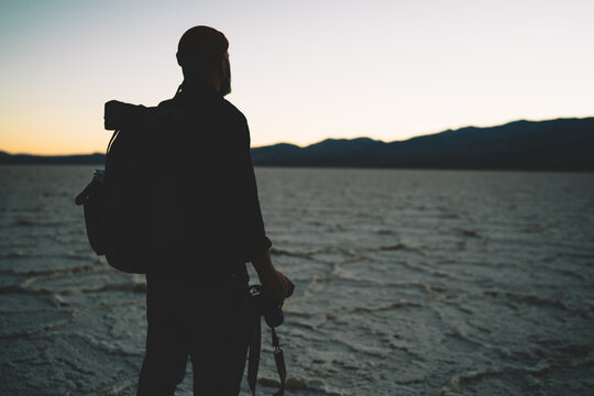 Male silhouette of traveler enjoying scenic skyline view on dawn in wild dry lake environment of Death valley, tourist standing with camera in Badwater basin during evening twiling after sunset .