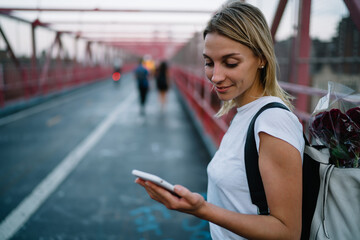 Young woman checking mail on smartphone while walking on urban settings carrying backpack with flowers,hipster girl browsing website and reading information via mobile phone while strolling on bridge.