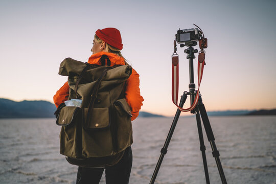 Back view of female photographer with backpack making preparation before starting to shoot video of scenic environment, woman taking photos of Badwater basin using modern equipment and tripod