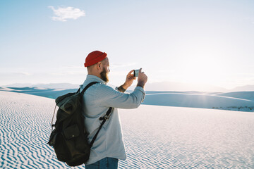 Trandy dressed hipster guy tourist making photo of beautiful sunset and landscape of bunes during hiking tour on vacations, male wanderlust influencer shooting video for sharing in travel blog