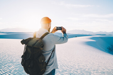 Obraz premium Back view of male traveler with rucksack making picture on smartphone camera exploring nature landmark in USA, guy wanderlust using app on telephone for shooting video from journey for blog