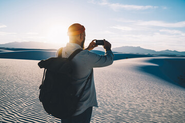 Back view of male blogger shooting video on smartphone during trip to White sands national park, guy wanderlust making picture of dunes on telephone exploiting wild nature with touristic backpack