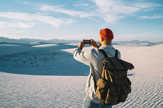 Guy wanderlust with backpack holding smartphone for taking beautiful picture of desert arid lands, male with touristic backpack standing with cellphone shooting video for travel vlog on trip.