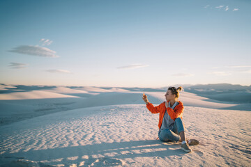 Young woman taking photo of beautiful nature background on trip to desert with White Sands in USA, female traveler sitting on dunes using telephone camera photographing scenery nature during vacations