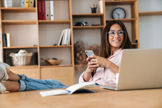 Image of smiling woman using cellphone while sitting with legs on table