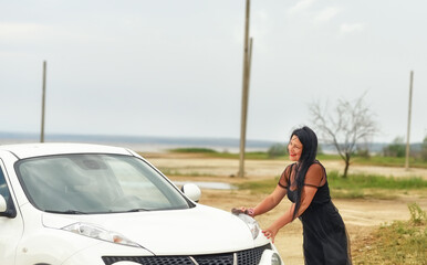 A girl with black hair in an excellent mood cheerfully wipes dust from a car in nature.