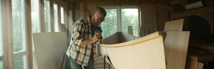 Mid 50s Caucasian male building a canoe in his workshop, using milling tool. Boat making hobby, small business owner