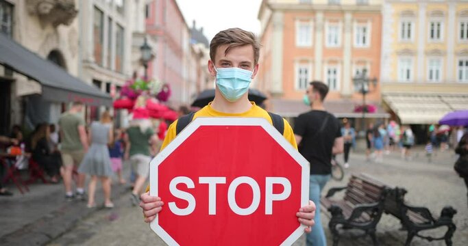A young man in a mask and with a stop sign in his hands stands on the street where many people do not comply with quarantine and social distance. Stop the coronavirus epidemic.
