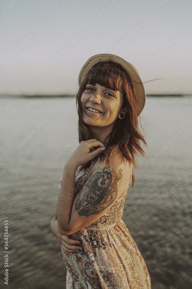 Wall mural young female with tattoos wearing a dress and straw hat on blurred ocean background