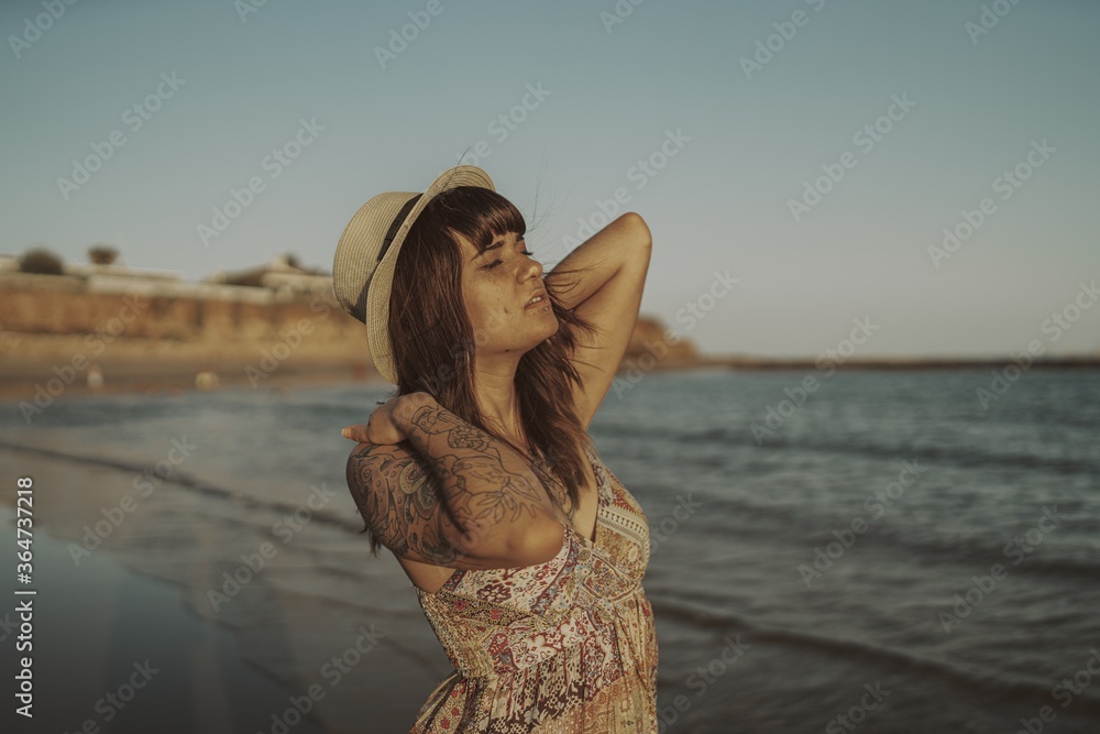 Sticker young female with tattoos wearing a dress and straw hat on blurred ocean background