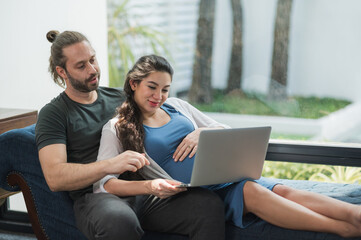 Beard man and his pregnant wife watching movie on laptop feeling happy while sitting on sofa in the...